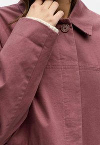 Brązowoczerwona marynarka z zapięciem na guziki, posiadająca spiczasty kołnierz i teksturowaną tkaninę. Rękaw podszyty kontrastowym jasnym materiałem.