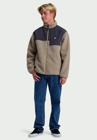 Fleece jacket in beige with a dark gray upper section; features a zip closure and a logo patch. Paired with blue jeans and black shoes.