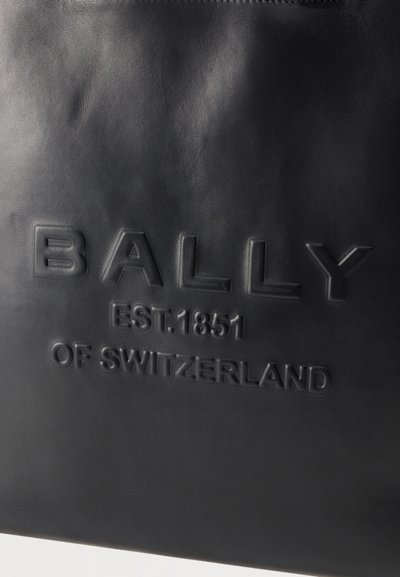 Surface en cuir noir embossée du texte "BALLY EST. 1851 DE SUISSE" en lettres majuscules.