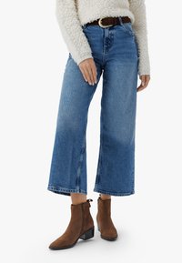 Kvinna klädd i blå vida jeans, en krämfärgad texturerad tröja, brunt bälte och bruna mockakängor på en vit bakgrund.
