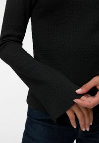 Czarna, teksturowana bluzka z długim rękawem i rozszerzanymi mankietami, z wyróżniającym się wzorem i dopasowanym krojem. Ręce spoczywają na mankietach.