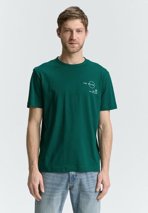 Zielony t-shirt z bawełny z krótkimi rękawami i okrągłym dekoltem. Posiada białe napisy graficzne na lewej stronie klatki piersiowej. Noszony z jasnoniebieskimi dżinsami.