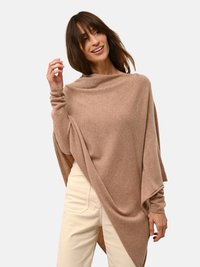 Maglione oversize marrone chiaro realizzato in un tessuto morbido, con un collo alto e un design drappeggiato, abbinato a pantaloni di colore chiaro.