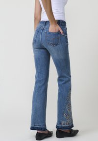 Jeans de mezclilla azul con corte recto, con bordados coloridos a lo largo de la parte inferior de la pierna y bolsillos traseros estándar. Se llevan con zapatos negros.