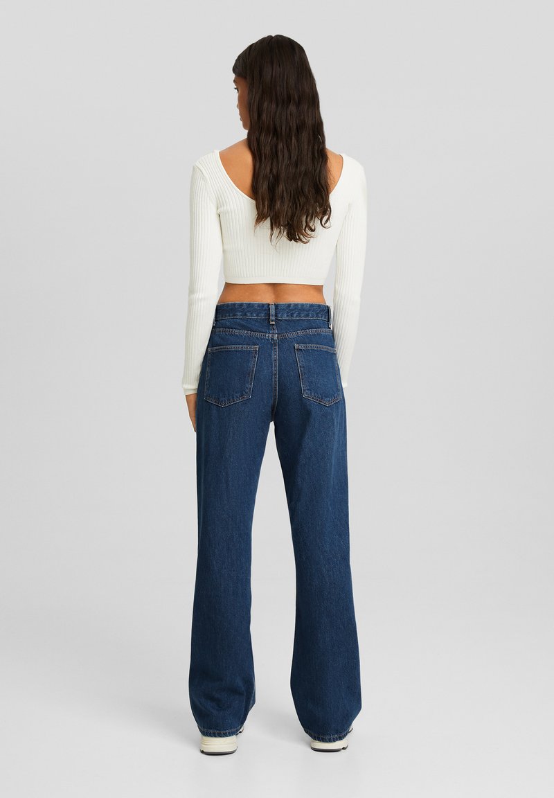 Un haut blanc à manches longues côtelé associé à un jean en denim bleu taille haute et à jambes larges. Le jean dispose de deux poches arrière et d'une coupe décontractée.