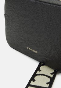 Borsa a tracolla nera in pelle con superficie testurizzata, logo dorato e una tracolla in tessuto a motivo crema e nero.