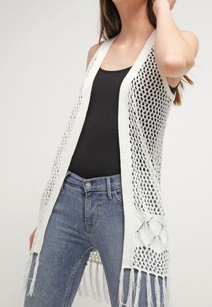 Femme portant un gilet blanc en crochet sans manches avec des franges par-dessus un top noir et un jean bleu.