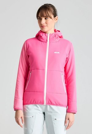 Una mujer luce una chaqueta con capucha de color rosa intenso y cremalleras blancas, con unos pantalones claros, sobre un fondo gris liso.