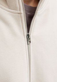 Zbliżenie na częściowo zasunięty srebrny metalowy suwak na beżowej kurtce z tkaniny, pod którą znajduje się brązowa koszula.