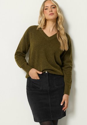 Femme aux longs cheveux blonds portant un pull à col en V vert olive et une jupe en velours côtelé noire, posant avec une main dans la poche devant un mur blanc.