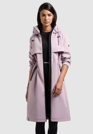 Donna che indossa un cappotto con cappuccio viola chiaro fino al ginocchio sopra abiti neri, in piedi contro uno sfondo chiaro e uniforme.