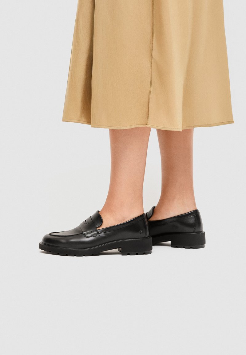 Μαύρα δερμάτινα μοκασίνια με χοντρές σόλες, φορεμένα με μπεζ midi φούστα, εμφανιζόμενα σε πόδια πάνω σε απλό ανοιχτό φόντο.