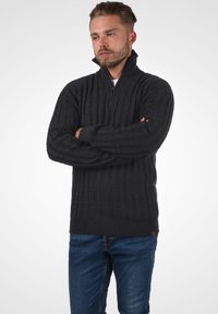 Dunkelgrauer Kabelstrickpullover mit einem halben Reißverschlusskragen und geripptem Saum. Kombiniert mit blauen Jeans, die ein strukturiertes Stoffdesign zeigen.