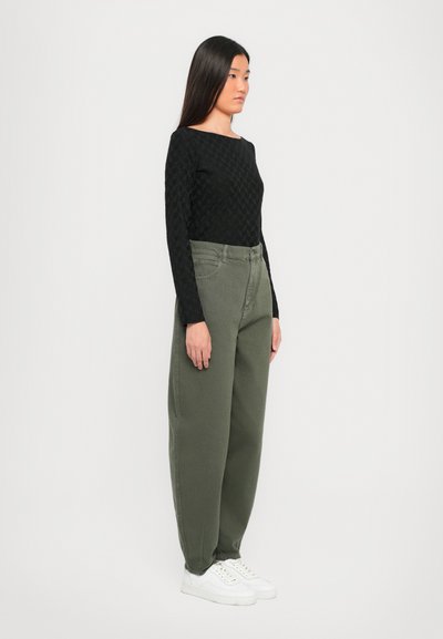 Maglietta nera a maniche lunghe con motivo testurizzato, abbinata a pantaloni larghi di colore verde oliva. Modella indossa sneakers bianche, in piedi su uno sfondo bianco.