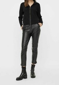 Pull à fermeture éclair noir en tissu doux, assorti à un pantalon en cuir noir ajusté avec une finition brillante et une coupe à la cheville, complété par des bottes noires épaisses.