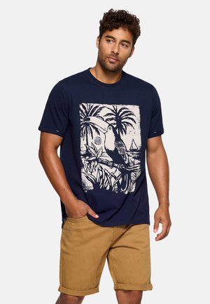 Hombre con camiseta azul marino con estampado de tucán tropical y palmera, combinada con pantalones cortos beige remangados, de pie con una mano en el bolsillo.