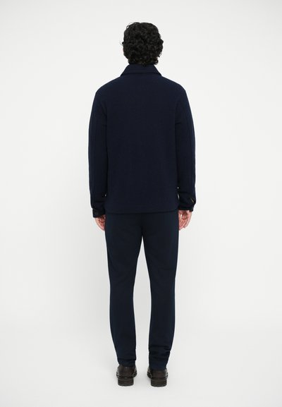 Veste en laine mélangée navy avec col et poignets à boutons, assortie à un pantalon navy et des chaussures sombres. Vue de dos.