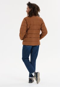 Brun pufferjakke med vandrette syninger, tætsiddende silhuet og to sidelommer. Båret over blå jeans og sorte sneakers.