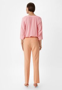 Rosa Bluse mit gerafftem Rücken, Dreiviertelärmel. Pfirsichfarbene, maßgeschneiderte Hose mit Gesäßtaschen. Sandalen mit Absatz in passender Farbe.