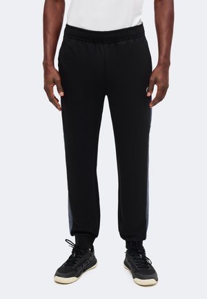 Mann trägt schwarze sportliche Jogginghosen mit elastischem Bund und schwarzen Sneakers, steht vor einem einfarbigen Hintergrund.