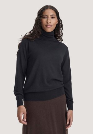 Femme aux longs cheveux bouclés portant un pull à col roulé noir et une jupe marron, debout devant un fond uni.