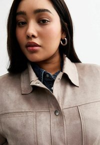 Chaqueta de ante de color taupe claro con cuello, botones a presión y una textura sutil, superpuesta sobre una camisa de mezclilla. Tonos tierra, diseño minimalista.