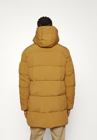 Pufferjas in een effen mosterdkleur, met een capuchon, horizontaal gekwilt patroon en een middellange snit met een soepele textuur.