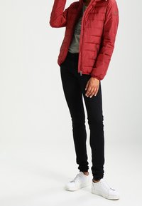 Osoba v červené prošívané bundě, šedém tričku s potiskem, černých slim kalhotách a bílých teniskách, stojící na jednobarevném pozadí.