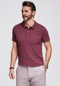 Polo shirt bordeaux a maniche corte, con colletto, patta con due bottoni e taschino sul lato sinistro del petto. Tessuto morbido, abbinato a pantaloni grigi chiari.