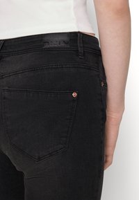 Melnas džinsi no denima ar gludu tekstūru, aizmugurējām kabatām ar šuvju detalizāciju un ādas zīmola uzlīmi uz jostasvietas.