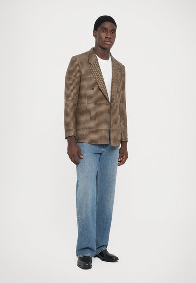 Brun rutmönstrad dubbelknäppt kavaj med slag, tillsammans med ljusblå vida jeans och svarta läderskor.