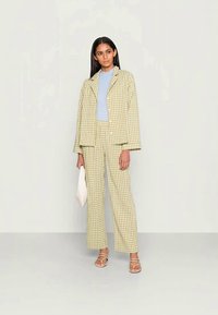 Conjunto de fato em padrão xadrez amarelo e verde, com um casaco de corte solto e calças de perna reta. Blusa azul clara por baixo, segurando uma bolsa de mão branca.