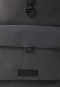Black waterproof backpack with a smooth texture, flap closure, and branded leather patch. Features sturdy buckle hardware and contrasting stitching.