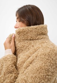Beige, fluffy jasje met hoge kraag gemaakt van textuurstof, met zichtbare stiksels. Het model houdt de kraag vast terwijl ze een zijprofiel toont.