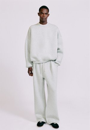 Jeune homme debout portant un sweat-shirt gris clair ample et un pantalon de jogging assorti avec des chaussures slip-on noires sur un fond uni.