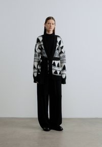 Πλεκτή ζακέτα σε ασπρόμαυρο με μοτίβο και ζώνη, που παρουσιάζει γεωμετρικό σχέδιο και μακριά μανίκια, σε συνδυασμό με φαρδιά, μαύρα παντελόνια.