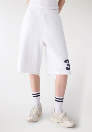 Persona che indossa pantaloncini bianchi fino al ginocchio con numero "3" blu navy sul lato, calzini bianchi con strisce nere e scarpe da ginnastica beige su sfondo neutro.