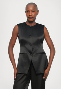 Sieviete ar noskūtu galvu, tērpusies bezmugurē melnā satīna žaketē un saskaņotās biksēs, stāv ar rokām atviegloti pie sāniem.