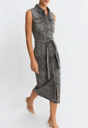 Abito midi in denim grigio lavato senza maniche con bottoni frontali, tasche sul petto, cintura in vita, spacco laterale, indossato con scarpe beige con tacco alto.