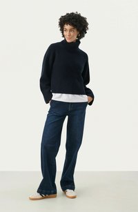 Dark navy ribbed sweater with a high collar, layered over a white shirt, paired with loose dark denim jeans and white sneakers.