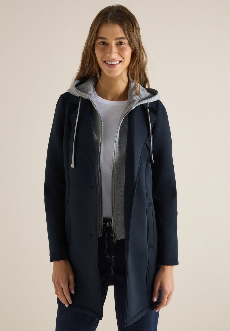 Jeune femme souriante, portant des vêtements superposés avec un sweat à capuche gris sous un manteau bleu marine foncé, une chemise blanche et un jean foncé, sur un fond uni.