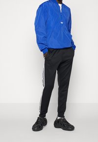 Modrá nylonová bunda s polovičním zipem a klokáním kapsou, doplněná černými tepláky s bílými bočními pruhy a černými sportovními botami.