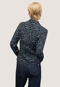 Upnutá černá blůza s modrým tečkovaným vzorem, dlouhými rukávy a límečkem. Viděná ze zadní strany v kombinaci s tmavými džínami.
