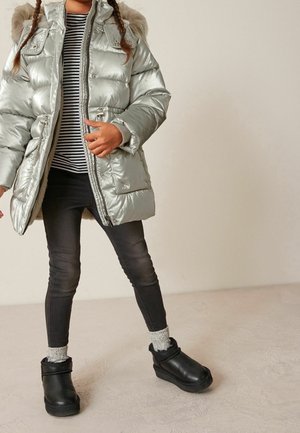 Enfant portant une veste matelassée argentée avec capuche en fourrure, des leggings noirs, un t-shirt rayé, des chaussettes grises et des bottines noires, debout sur un sol clair.