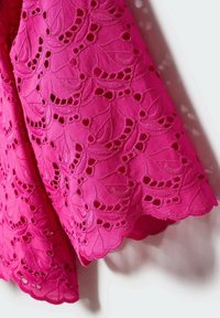 Tissu rose vif avec des motifs floraux brodés et de petites découpes circulaires, détail d'ourlet festonné visible sur un fond clair.