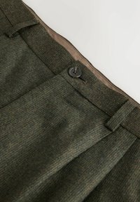Dunkelgrüne Tweed-Hosen mit einem Knopfverschluss, Falten und einem glatten beigen Innenfutter, das am Bund sichtbar ist.