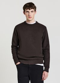 Brauner Strickpullover mit Rundhalsausschnitt, gerippten Bündchen und Saum. Wird über einem weißen Hemd getragen und zeichnet sich durch einen strukturierten Stoff in entspannter Passform aus.