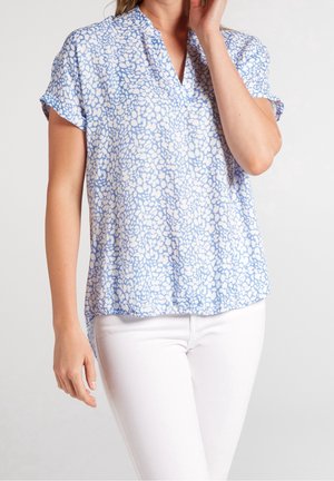 Donna con una camicetta a maniche corte azzurro chiaro con motivi floreali bianchi abbinata a pantaloni bianchi, in piedi davanti a uno sfondo semplice.