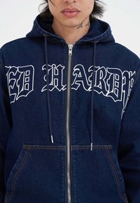 Sudadera con cremallera de mezclilla azul oscuro que presenta texto bordado en blanco en la parte frontal, dos bolsillos laterales y cordones en la capucha.