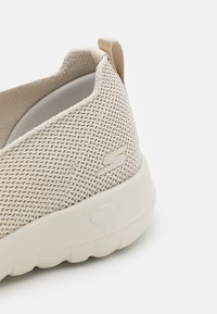 Chaussure slip-on en tricot beige avec une finition texturée, intérieur rembourré et semelle légère. Présente un logo discret sur le côté et l'onglet arrière.